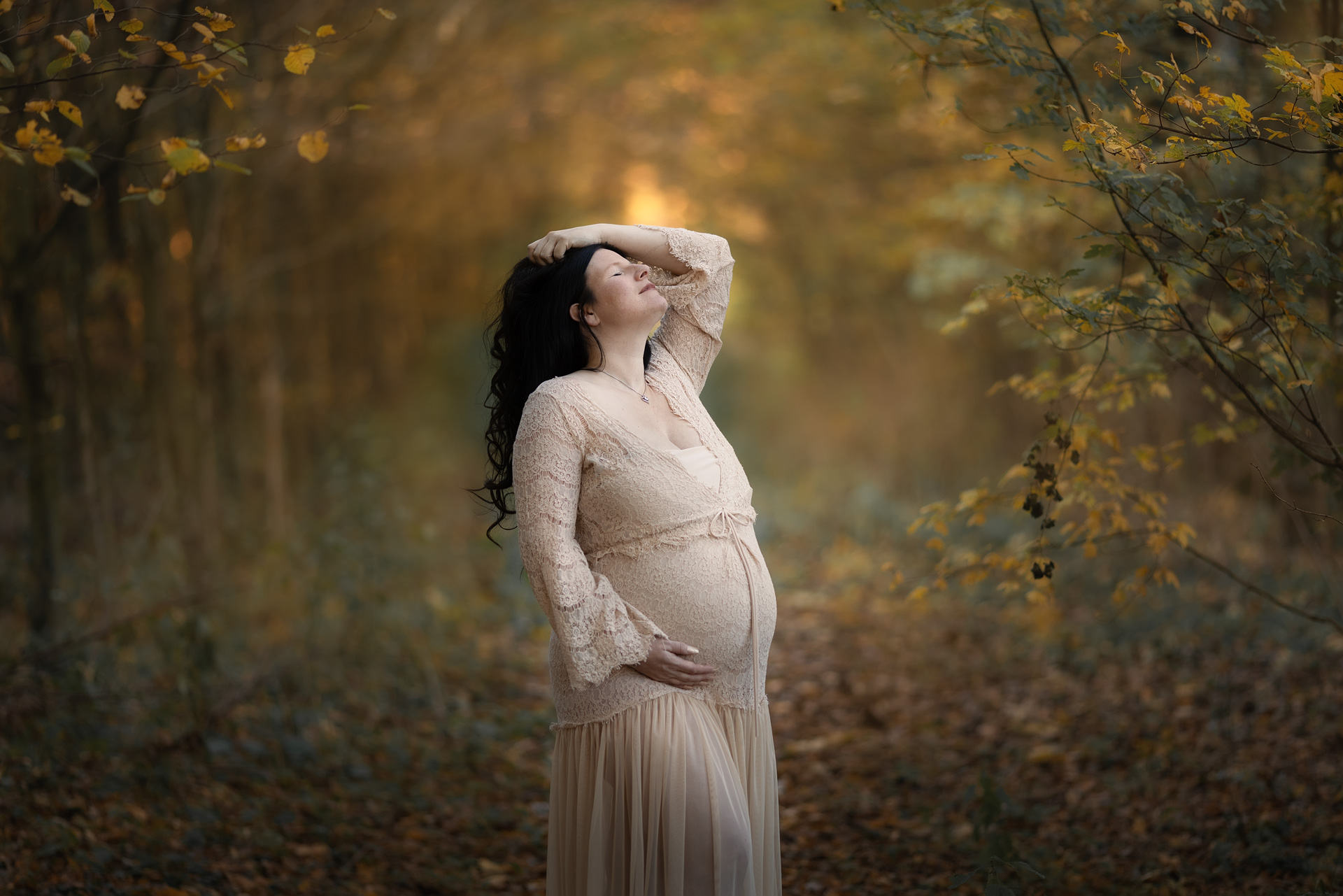 Babybauchfotografie im Wald. Frau mit dunklen Haaren und beigem Schwangerschaftskleid