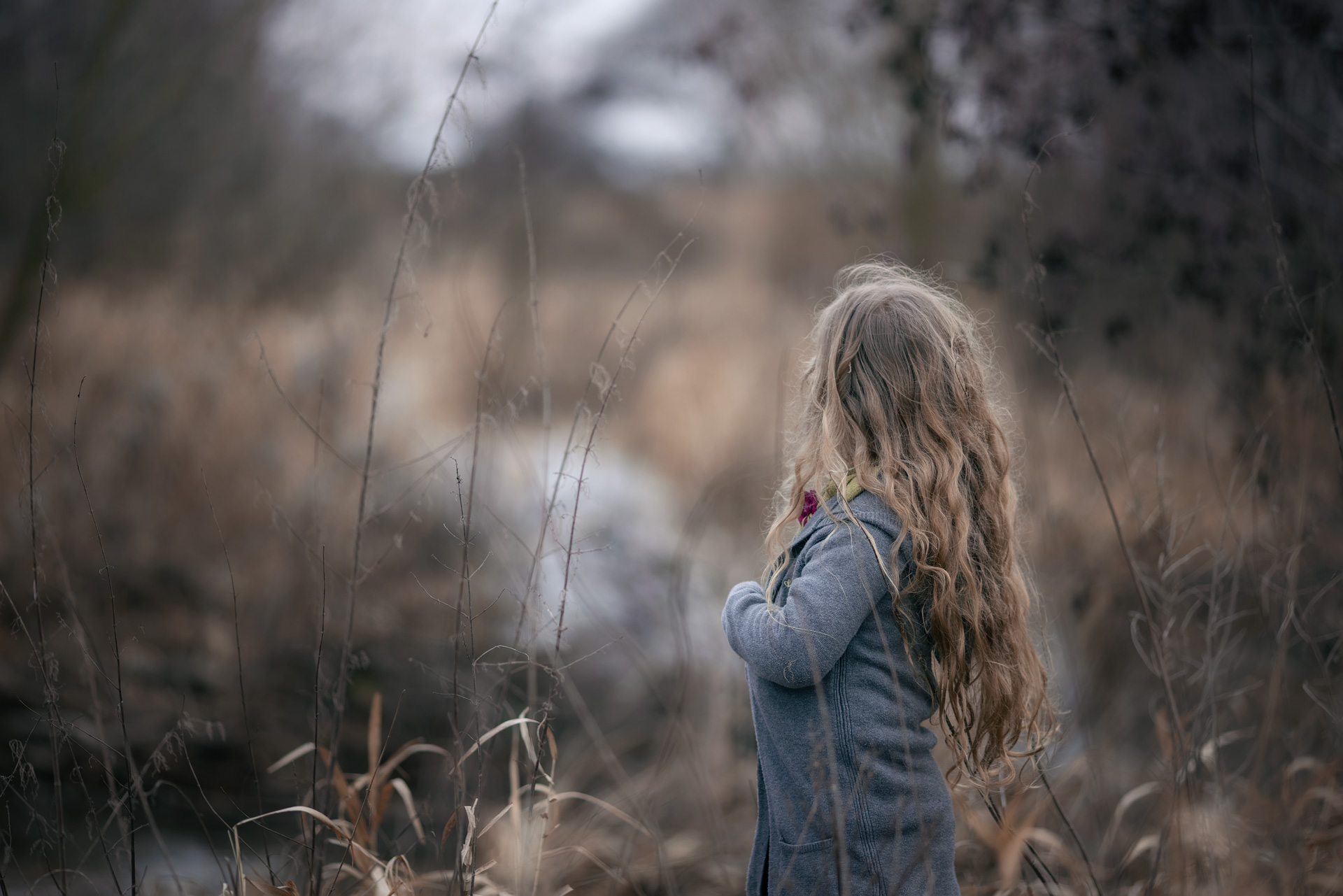 Kindershooting am Bach 2-Yvonne Pesch Fotografie Mädchen in grauem Strickmantel steht im alten hohen Gras und schaut in die Ferne. das Gesicht ist abgewand