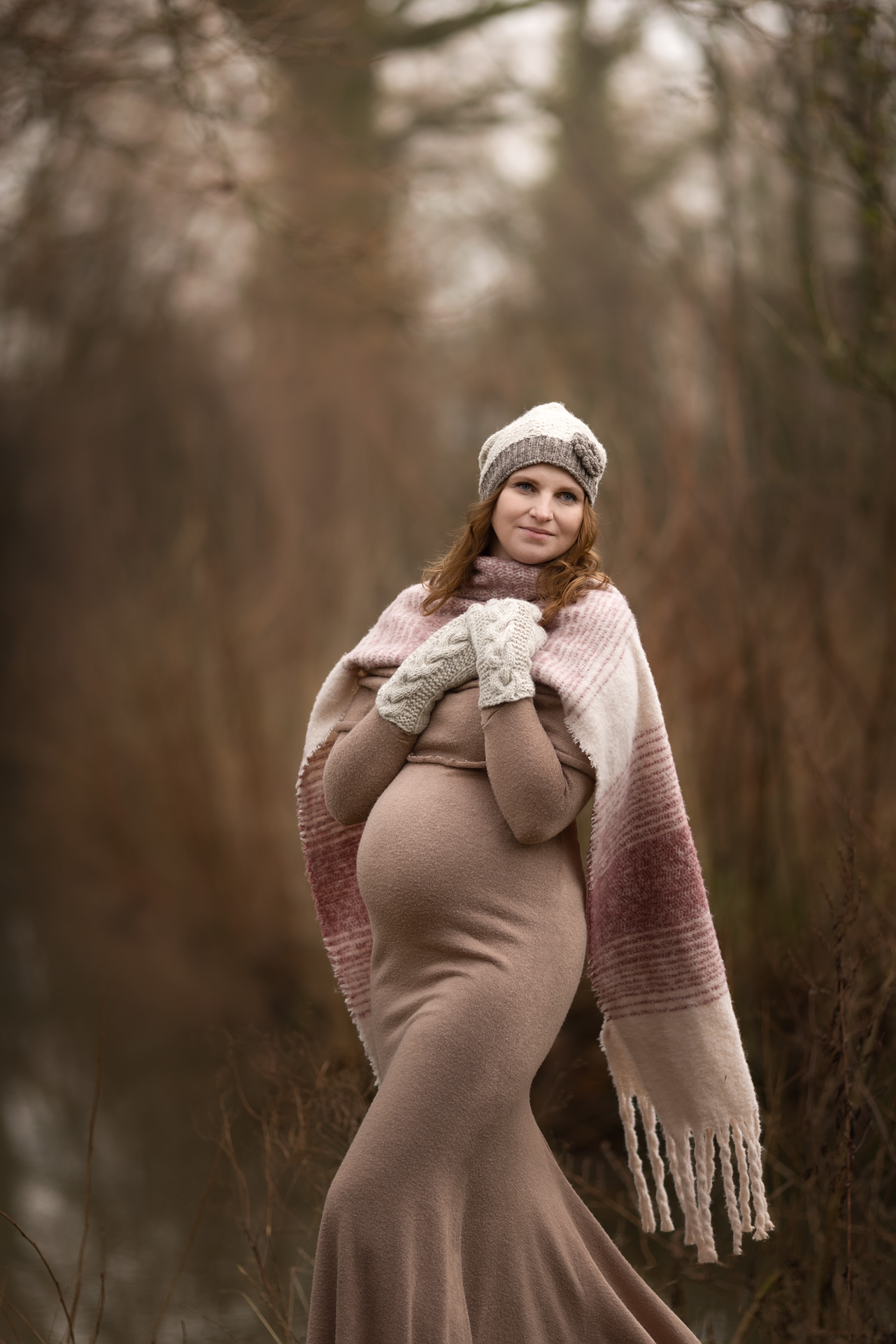 Babybauchfotografie im beigen Kleid mit Wollmütze und Schal in Winterlandschaft