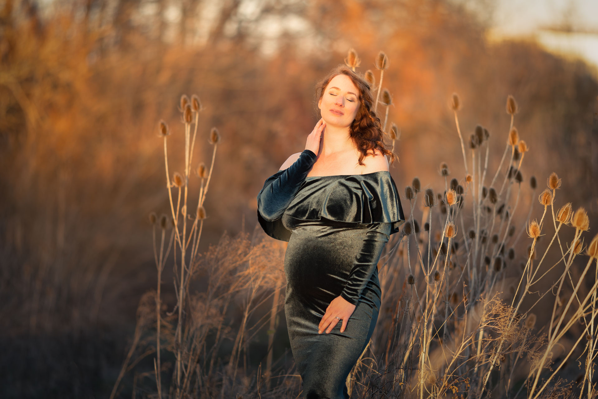 schwangere Frau im petrolfarbenen Samtkleid steht zwischen Disteln im hohen trockenen Gras