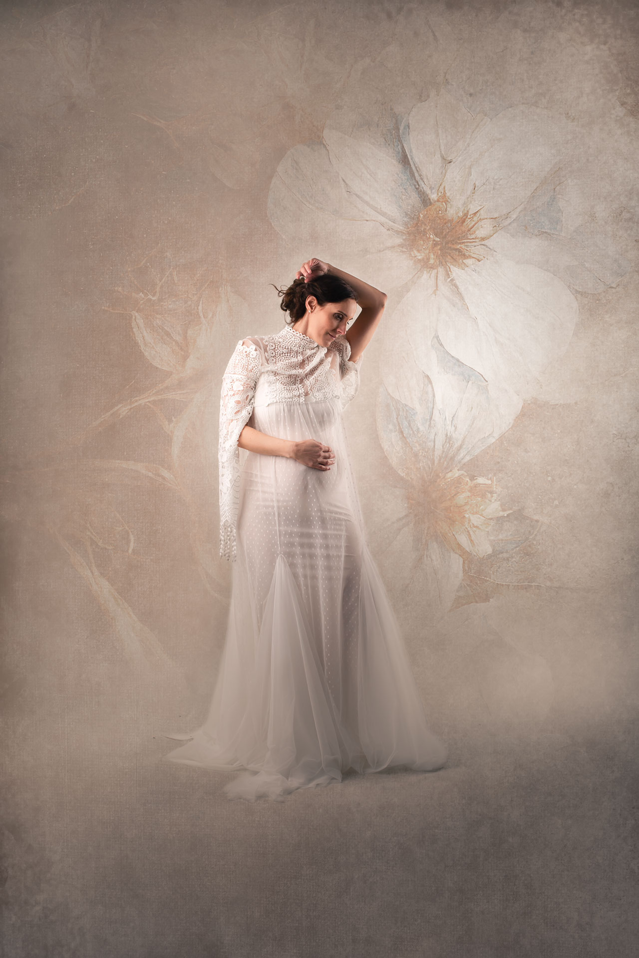 Schwangere Frau im weißen Kleid steht im Studio vor blumigem Hintergrund