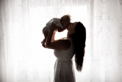 Mutter küsst ihr Baby aud die Stirn, welches sie vor sich hält. Sie steht am Fenster.