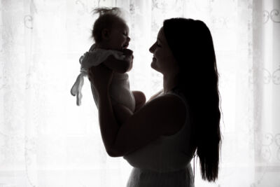 Gegenlichtaufnahme vorm Fenster. Mutter hält ihr Baby hoch