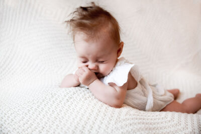 Baby liegt auf dem Bauch auf einer weißen Decke und kneift die Augen zu. Es hat eien weißen Body an