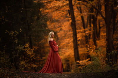 Babybauchshooting mit rotem Shootingkleid im Wald Schwangere blonde Frau im roten Kleid mit Schleppe steht auf einem Waldweg und hält ihren Bauch. Sie schaut in die Ferne