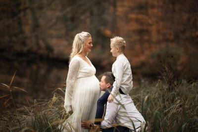 Schwangerschaftsshooting in der Natur Schwangere Frau im weißen Kleid schaut ihren Sohn an. Der Sohn sitzt auf den Schultern des Vaters. Der Vater hockt vor seiner Frau und küsst ihren Bauch.