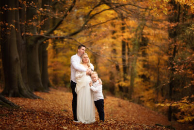 Familienbild mit schwangerer Frau im weißen Kleid unter Bäumen Mann umarmt von hinten seine schwangere Frau. Sie stehen im Wald. Der Sohn lehnt am Bauch der Mutter