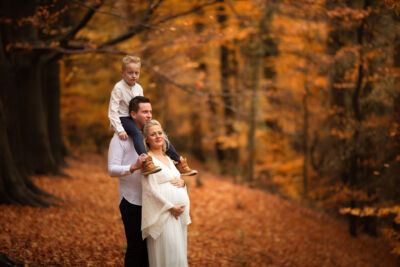 Familienbild mit schgwangerer Frau im weißen Kleid im Wald Schwangere Frau im weißen Kleid lehnt sich an ihren Mann. Sie stehen im herbstlichen Wald. Der Mann hat den Sohn auf den Schultern sitzen