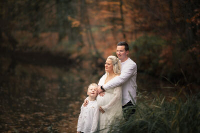 Familienbild mit schwangerer Frau im weißen Kleid schwangere Frau steht im weißen Kleid am Wasser. Vor ihr steht ihr kleiner Sohn im weißen Hemd und hinter ihr der Mann im weißen Hemd