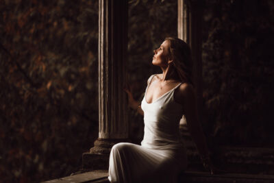 Frau im weißen Kleid sitzt an einer Säule und schaut in die Ferne