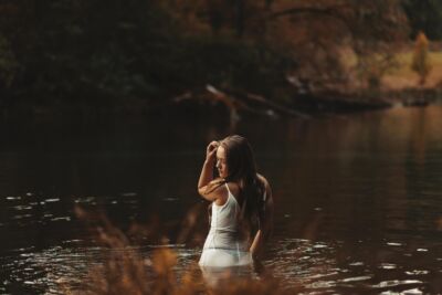 Frau im weißen Kleid steht mit dem Rücken zum Betrachter im See und hält die linke Hand am Kopf. Der Blick ist nach links gerichtet.