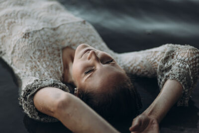 Frau im weißen Häkelkleid liegt im Wasser und streckt die Arme nach hinten