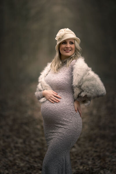 Babybauchfoto im grauen Spitzenkleid und Fellstola mit weißer Filzmütze im Wald