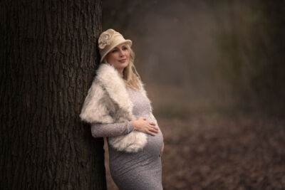 Babybauchfoto im grauen Kleid, Hut und Fellstola, angelehnt an einem Baum